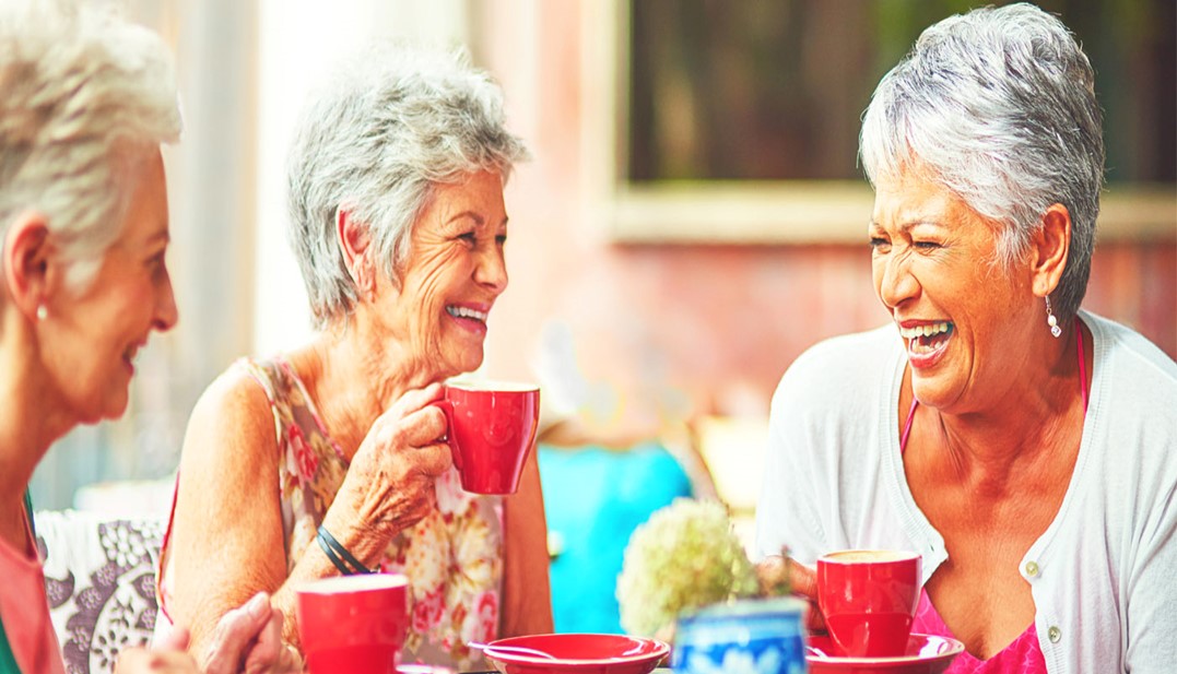 Três mulheres idosas conversando e sorrindo com uma xícara de café nas mãos.